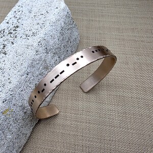 May include: A copper-coloured cuff bracelet featuring Morse code symbols. The open-ended bracelet sits on a textured, neutral-toned surface. The Morse code is represented by a series of dots and dashes.