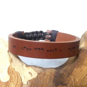 May include: A brown leather bracelet with a black braided cord closure. The bracelet has a message in braille embossed on the leather.