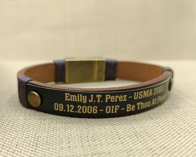 Puede incluir: Un brazalete de cuero marr&oacute;n con una banda negra grabada con el texto "Emily J.T. Perez - USMA 2005 09.12.2006 - OIF - Be Thou At Peace".