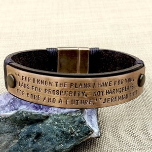 May include: A brown leather bracelet with a copper-colored metal plate. The plate has the text "FOR I KNOW THE PLANS I HAVE FOR YOU, PLANS FOR PROSPERITY, NOT HARM; PLANS FOR HOPE AND A FUTURE." JEREMIAH 29:11.