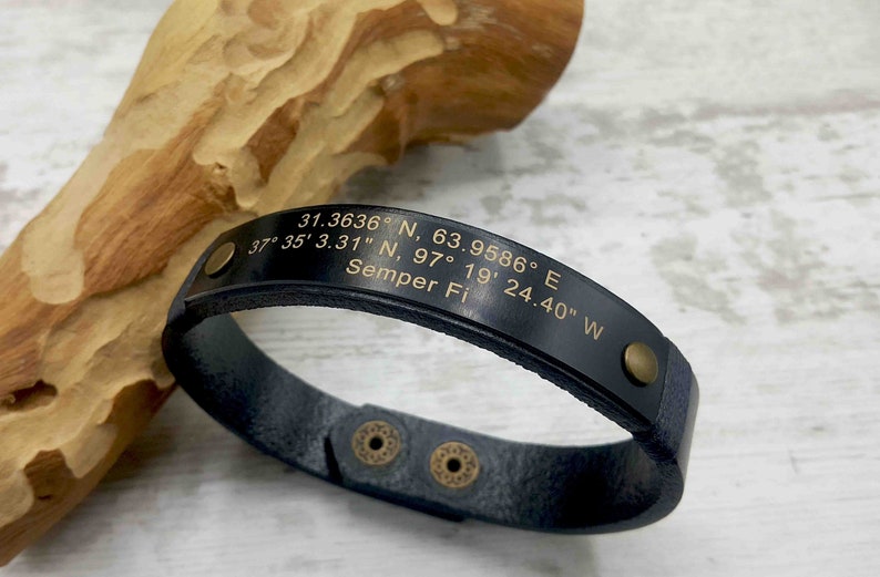 May include: Black leather bracelet with gold-tone accents and engraved coordinates and the text "Semper Fi".
