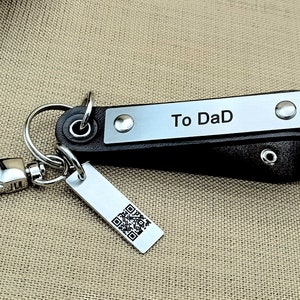 Könnte beinhalten: Schwarzer Leder-Schlüsselanhänger mit einer silbernen Platte mit der Aufschrift "To DaD". Er hat einen silbernen Karabinerverschluss und ein kleines rechteckiges Etikett mit einem QR-Code. Der Hintergrund ist ein beiger Stoff.