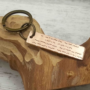 May include: A copper-colored rectangular keychain with Morse code text and a bronze-colored key ring. The keychain is attached to a bronze-colored key ring. The Morse code is etched into the metal. The keychain is displayed on a wooden surface.