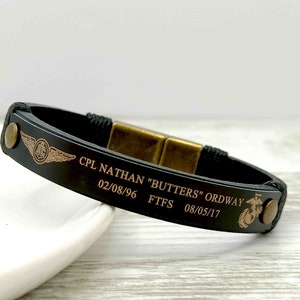 Puede incluir: Pulsera de cuero negro con cierre dorado y texto grabado: "CPL NATHAN "BUTTERS" ORDWAY 02/08/96 FTFS 08/05/17". La pulsera tambi&eacute;n presenta una insignia militar dorada.