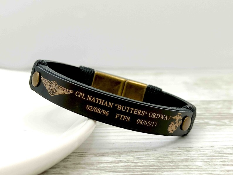 Puede incluir: Pulsera de cuero negro con cierre dorado y texto grabado: "CPL NATHAN "BUTTERS" ORDWAY 02/08/96 FTFS 08/05/17". La pulsera tambi&eacute;n presenta una insignia militar dorada.