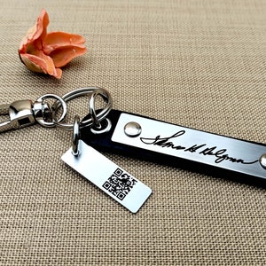 Könnte beinhalten: Ein schwarzer Leder-Schlüsselanhänger mit einem silbernen Metalletikett, auf dem die Unterschrift "Sarah McLachlan" eingraviert ist. Der Schlüsselanhänger hat außerdem ein silbernes Metalletikett mit einem QR-Code.
