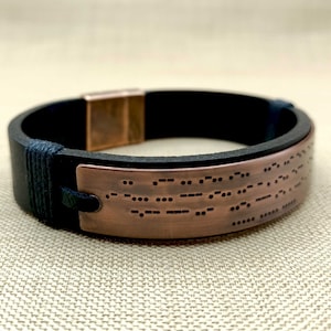 May include: A black leather bracelet with a copper plate engraved with braille. The bracelet has a magnetic clasp.