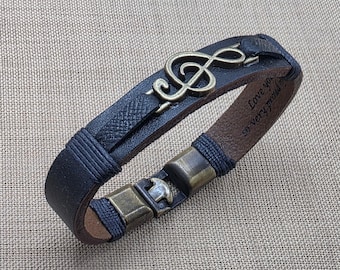 May include: Black leather bracelet featuring a treble clef charm. The bracelet has a textured exterior and a brown interior. The clasp is a bronze color. The text "Love you, so very proud" is engraved on the inside of the bracelet.