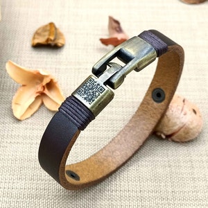 May include: A brown leather bracelet with a metal clasp and a black braided cord. The clasp has a QR code design.