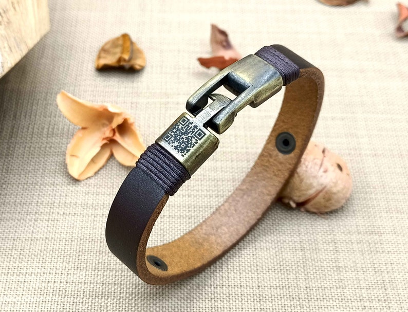 May include: A brown leather bracelet with a metal clasp and a black braided cord. The clasp has a QR code design.