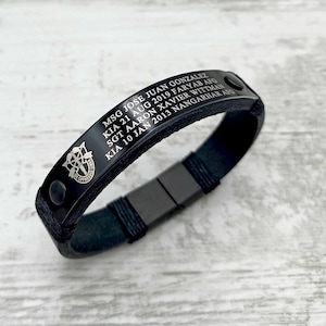 Könnte beinhalten: Ein schwarzes Lederarmband mit einer zentralen Metallplatte, die mit dem Text MSG JOSE JUAN GONZALEZ KIA 21 AUG 2019 FARYAB AFG SGT AARON XAVIER WITTMAN KIA 10 JAN 2013 NANGARHAR AFG und einem militärischen Abzeichen graviert ist. Mit schwarzem Metallverschluss.