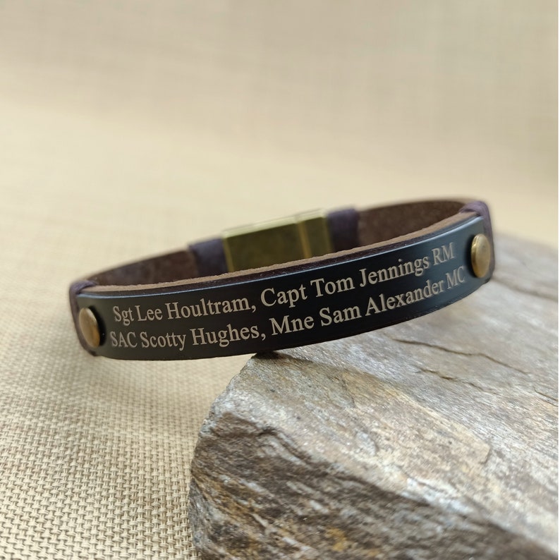 Puede incluir: Un brazalete de cuero marr&oacute;n con cierre magn&eacute;tico. El brazalete est&aacute; grabado con el texto "Sgt Lee Houltram, Capt Tom Jennings RM SAC Scotty Hughes, Mne Sam Alexander MC".