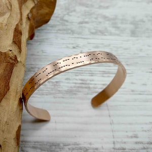 May include: A copper-colored cuff bracelet with a hammered texture. The bracelet is engraved with Morse code. The open cuff design allows for easy wear. The bracelet is displayed on a light wood surface.