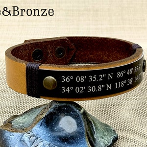 May include: A brown leather bracelet with a black leather strap and a metal rivet. The bracelet has two sets of coordinates engraved on it: 36&deg;08' 35.2" N 86&deg; 48' 55.9 and 34&deg; 02' 30.8" N 118&deg; 38' 14.0".