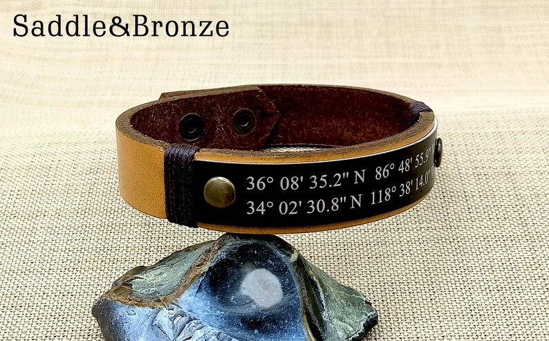 May include: A brown leather bracelet with a black leather strap and a metal rivet. The bracelet has two sets of coordinates engraved on it: 36&deg;08' 35.2" N 86&deg; 48' 55.9 and 34&deg; 02' 30.8" N 118&deg; 38' 14.0".