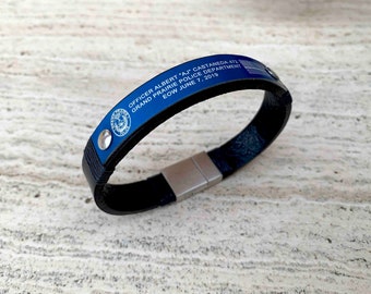 May include: A black leather bracelet with a blue metal plate. The plate has the text "OFFICER ALBERT 'AJ' CASTANEDA 472 GRAND PRAIRIE POLICE DEPARTMENT EOW JUNE 7, 2019" and a police badge. The bracelet has a magnetic clasp.