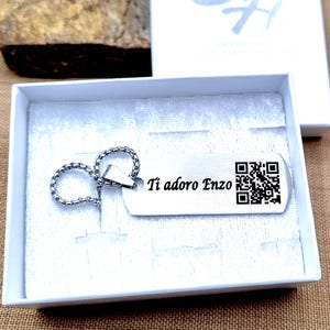 Peut inclure: Un porte-clés argenté avec une étiquette rectangulaire à l'intérieur d'une boîte blanche. L'étiquette porte le texte "Ti adoro Enzo" et un code QR. Le porte-clés est attaché à un anneau de type chaîne.