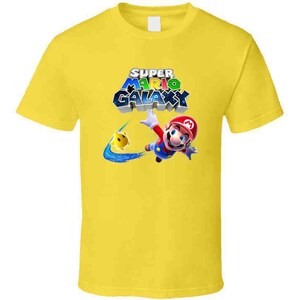 May include: Yellow t-shirt with a graphic of Mario from Super Mario Galaxy. The graphic shows Mario in a red and blue suit, flying through the air with a yellow star. The text "Super Mario Galaxy" is written in colorful letters above Mario.