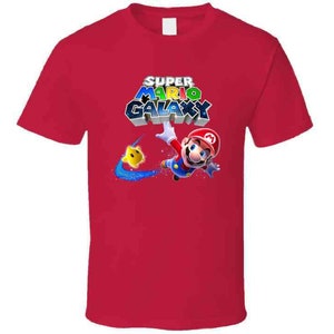 May include: Red t-shirt with a graphic of Super Mario Galaxy. The graphic features Mario in a blue and red suit, flying through space with a yellow star.