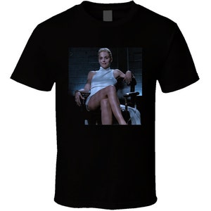 May include: Black t-shirt featuring a movie still of a woman in a white dress sitting in a chair. The image is a close-up, showing the woman's face and upper body. The background is a dark, industrial setting.