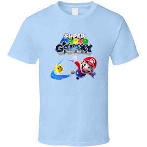 May include: Light blue t-shirt with a graphic of Mario from Super Mario Galaxy. The graphic features Mario in a red and blue suit, flying through the air with a yellow star. The text "Super Mario Galaxy" is printed in colorful letters above Mario.
