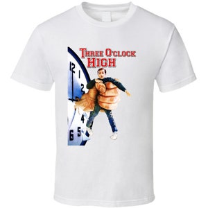 May include: White t-shirt with a graphic of a clock face and a man being held by a giant hand. The text "Three O'Clock High" is printed in red above the graphic.