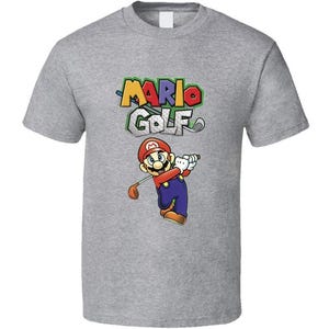 May include: A grey t-shirt with the "Mario Golf" logo and an illustration of Mario playing golf. The logo is in vibrant colours, and Mario is wearing his iconic red cap and blue overalls. A casual wear item.