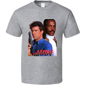 May include: A heather gray t-shirt featuring a movie poster graphic. The graphic includes two men, one holding a gun, with the text "LETHAL WEAPON" in red and blue. The shirt is a classic crew neck style.