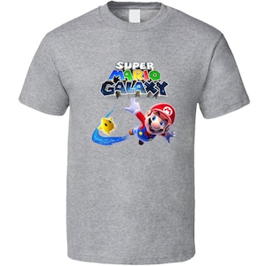May include: A gray t-shirt with a graphic of Mario from Super Mario Galaxy. The graphic shows Mario flying through space with a yellow star. The text "Super Mario Galaxy" is printed above Mario.