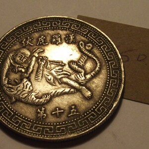 Peut inclure: Une pièce de monnaie chinoise en argent avec un motif de tigre et d'une personne. La pièce a une bordure de motifs géométriques répétés et le texte "火虎 肆 錢" et "寶 十 五" en caractères chinois.