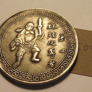 Peut inclure: Une pièce de monnaie chinoise en argent avec un motif en relief d'un homme tenant un bâton. La pièce a une bordure décorative avec un motif répétitif. Les caractères chinois sur la pièce de monnaie se lisent "Dao De Jing" qui se traduit par "Tao Te Ching".