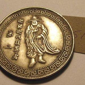 May include: Silver coin with a Chinese design featuring a man in a robe and hat. The coin has a border of repeating patterns.