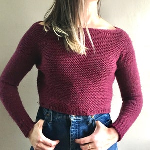 May include: A maroon knitted sweater with a wide neckline and long sleeves. The sweater is cropped and has a relaxed fit.