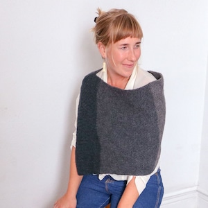 May include: A woman wearing a gray knitted poncho with a white shirt underneath. She is wearing blue jeans and has her hair pulled back.