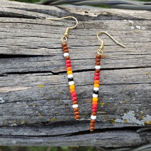 May include: A pair of long, beaded earrings with a brown, red, orange, white, and black color scheme. The earrings are hanging from gold hooks.