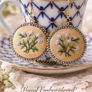 Op de afbeelding: Handgeborduurde bloemen oorbellen. Elke oorbel heeft een ronde, bruine stoffen basis met een bloemenontwerp in groene en lichtblauwe draad. De randen zijn afgewerkt met blauwe kralen en goudkleurig metaal. De oorbellen worden gepresenteerd op een schotel en voor een kopje.