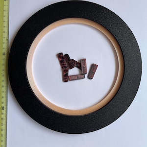 Pode incluir: Uma moldura circular preta com um anel interno castanho claro sobre um fundo branco. Vários objetos pequenos, retangulares, vermelho escuro com uma superfície estampada estão espalhados dentro da moldura. Uma régua é visível, medindo até 30 cm.