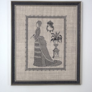 May include: A framed black and white cross-stitch embroidery depicting a woman in a long dress standing next to a potted plant. The embroidery is on a beige fabric and is framed in a silver frame.