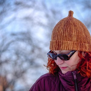 May include: A person wearing a brown knitted beanie with a small top detail and black sunglasses. The person is also wearing a purple jacket. The background is blurred with a mix of white and blue.