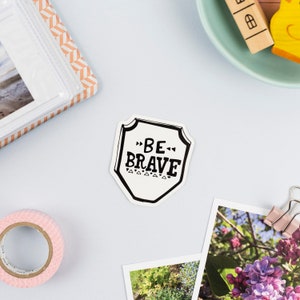 Puede incluir: Pegatina en blanco y negro con forma de escudo y el texto "BE BRAVE" en su interior.