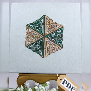 May include: A geometric cross-stitch design featuring a Celtic knot pattern in shades of green, peach, and gold. The artwork is displayed on a light blue canvas. A tag with the text "PDF" hangs from the canvas.