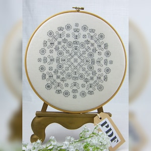 May include: A white embroidery hoop with a black and white geometric design. The hoop is on a wooden stand with a label that says "PDF".