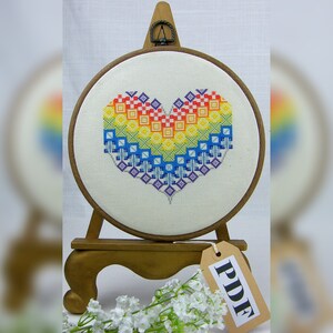 May include: A rainbow heart cross-stitch pattern is displayed in a wooden embroidery hoop on a stand. The hoop is decorated with a small wooden tag that reads "PDF".