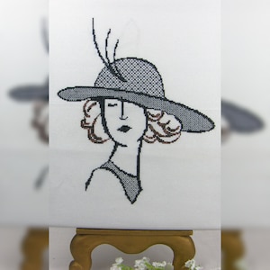May include: Black and white cross-stitch embroidery of a woman wearing a wide-brimmed hat with a feather detail.
