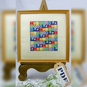 May include: A framed cross-stitch design with a colorful geometric pattern in shades of red, blue, green, yellow, and purple. The frame is made of wood and has a light brown finish. The cross-stitch is displayed on a wooden easel with a brown tag that says "PDF".
