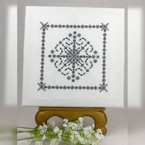 Peut inclure: Une toile blanche avec un motif géométrique brodé en noir. La toile est sur un chevalet doré et un bouquet de fleurs blanches est au premier plan.