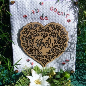Pode incluir: Um tecido branco com bordado de ponto cruz que l&ecirc; "Au Coeur de Noel" em franc&ecirc;s. Um recorte em forma de cora&ccedil;&atilde;o marrom com um design de filigrana preta est&aacute; centralizado no tecido.