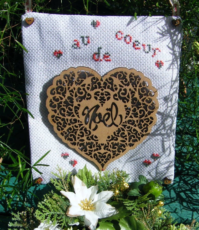 Pode incluir: Um tecido branco com bordado de ponto cruz que l&ecirc; "Au Coeur de Noel" em franc&ecirc;s. Um recorte em forma de cora&ccedil;&atilde;o marrom com um design de filigrana preta est&aacute; centralizado no tecido.