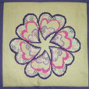 May include: A beige fabric pillow with a purple border features a cross-stitch design of a flower with multiple layers of hearts in shades of pink, purple, and green.