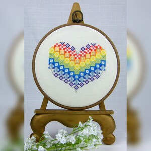 May include: A rainbow heart cross-stitch pattern is displayed in a wooden embroidery hoop on a gold stand. The hoop is surrounded by white flowers.